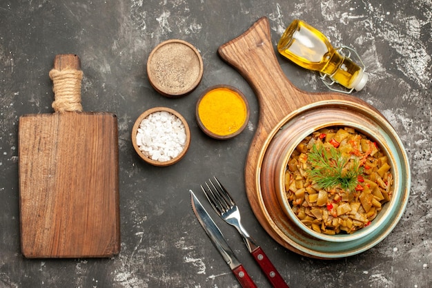 Vista dall'alto gustoso piatto di fagiolini con pomodori sul tabellone accanto alla forchetta coltello bottiglia di olio e tre tipi di spezie sul tavolo scuro