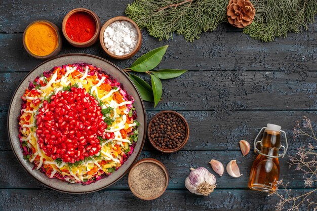 Top view tasty food appetizing Christmas food garlic bottle of oil and six bowls of colorful spices next to the spruce branches with cones