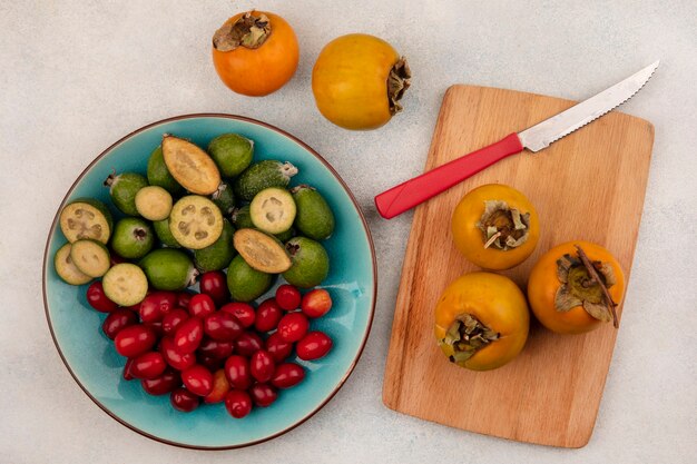 Top view of tasty fresh feijoas with cornelian cherries on a blue dish with persimmons on a wooden kitchen board with knife on a grey wall