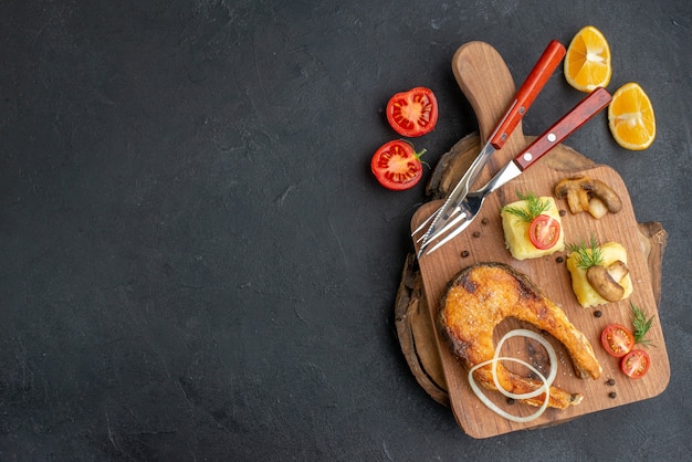 Top view of tasty fried fish and mushrooms tomatoes greens on cutting board cutlery set pepper on black surface