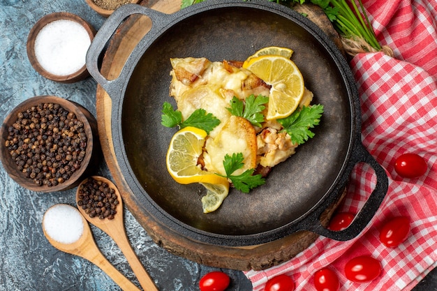 Top view tasty fried fish in pan on round wood board cherry tomatoes spices bowls wooden spoons on grey background