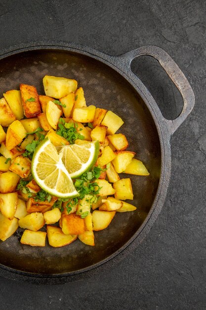 Вид сверху вкусного жареного картофеля внутри сковороды с ломтиками лимона на темной поверхности