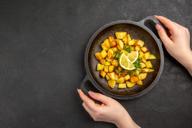 Вид сверху вкусного жареного картофеля внутри сковороды с ломтиками лимона на темной поверхности