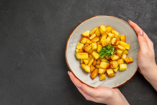 Вид сверху вкусного жареного картофеля внутри тарелки с зеленью на темной поверхности