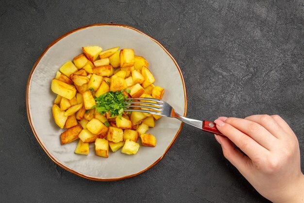 Top view of tasty fried potatoes inside plate with greens on dark surface