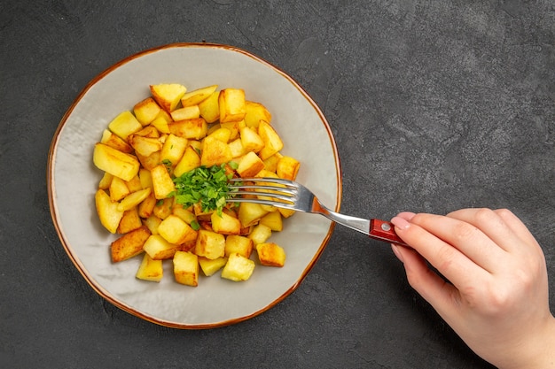 Top view of tasty fried potatoes inside plate with greens on dark surface