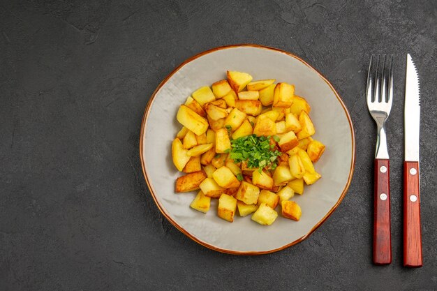 Top view of tasty fried potatoes inside plate with greens on dark surface