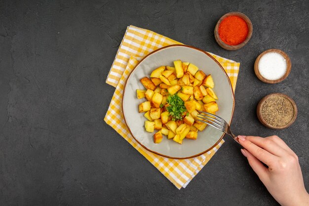 Вид сверху вкусного жареного картофеля внутри тарелки с приправами на темно-серой поверхности