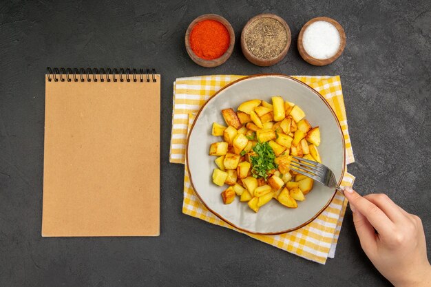 Top view of tasty fried potatoes inside plate with seasonings on a dark surface