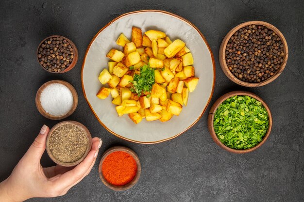 Top view of tasty fried potatoes inside plate with seasonings on dark surface