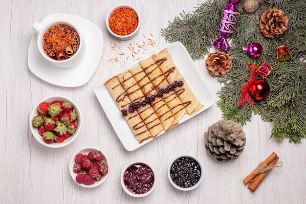 Top view of tasty pancake rolls with cup of tea and fruits on white