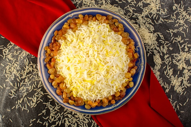 A top view tasty pilaf with oil and dried raisins inside plate with raw rice on the dark surface