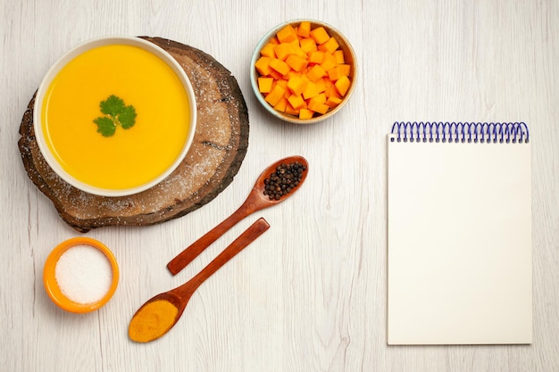 Top view of tasty pumpkin soup with seasonings and notepad on white