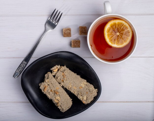 Top view of tasty slices of halva in a plate and a cup of tea with lemon slice on white