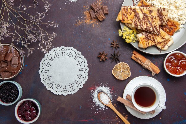 Вид сверху вкусной сладкой выпечки с чашкой чая и джемом на темном столе, печенье, бисквитный торт, сахар, сладкий чай