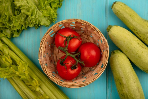 Top view of tasty tomatoes on a bucket with lettuce zucchinis and celery isolated on a blue wooden surface