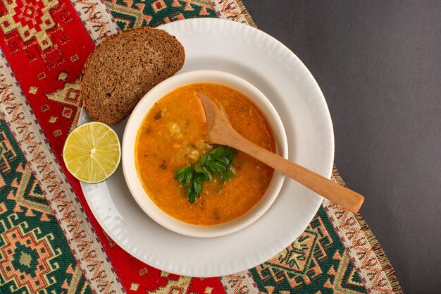 Вид сверху вкусного овощного супа внутри тарелки с буханкой хлеба и лимоном на темной поверхности