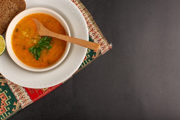 Top view of tasty vegetable soup inside plate with bread loaf and lemon on the dark surface