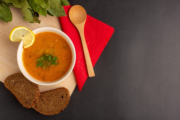 Top view of tasty vegetable soup inside plate with lemon slice and bread loafs on the dark surface