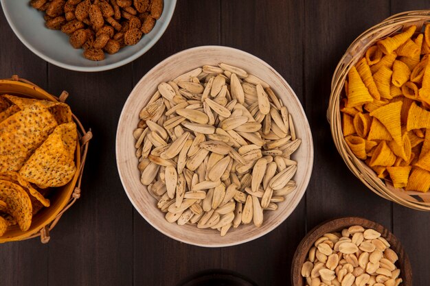 Top view of tasty white sunflower seeds on a bowl with spicy chips on a bucket with corn snacks on a bucket on a wooden table