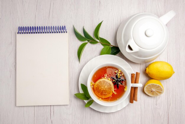 Top view tea and cinnamon a cup of herbal tea cinnamon sticks on the saucer next to the white notebook lemon teapot and leaves on the white table