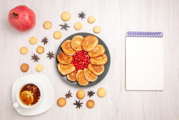 テーブルの上のハーブティーザクロとクッキーのカップザクロとパンケーキの暗いプレートの横にある上面図のお茶とパンケーキの白いノート