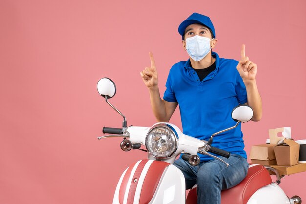 Top view of thinking courier guy in medical mask wearing hat sitting on scooter delivering orders and pointing up on pastel peach