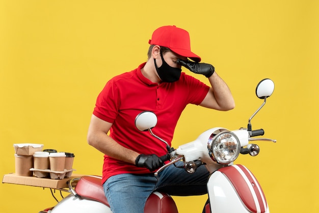 Foto gratuita vista dall'alto del giovane adulto di pensiero che indossa guanti di camicetta e cappello rossi nella mascherina medica che trasporta l'ordine che si siede sul motorino sulla parete gialla