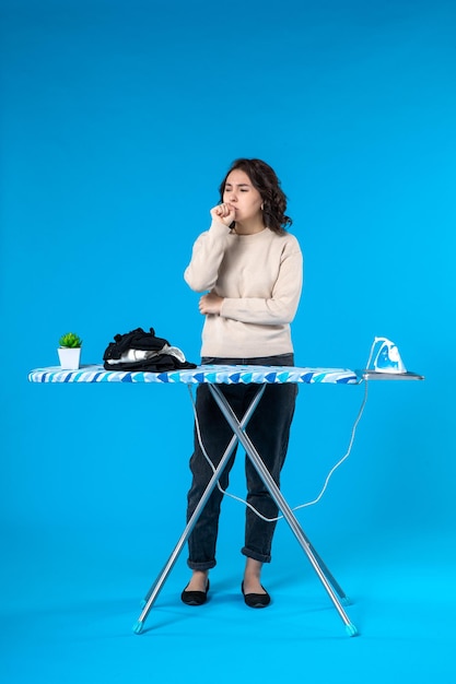 Top view of thinking young girl standing behind the ironing board and thinking deeply on blue wave background