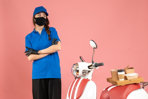 Vista dall'alto della premurosa ragazza del corriere in maschera medica in piedi accanto alla moto con una torta di caffè su di essa su color pesca pastello