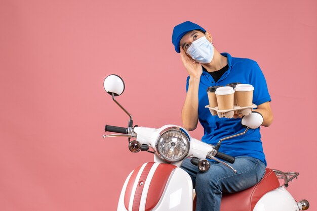 Top view of thoughtful courier man in mask wearing hat sitting on scooter showing orders on pastel peach