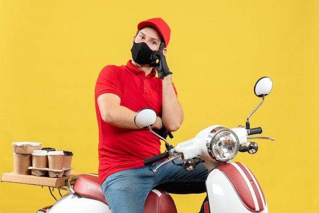 Top view of thoughtful young adult wearing red blouse and hat gloves in medical mask delivering order sitting on scooter on yellow wall