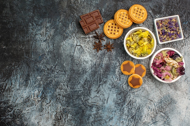 Top view of three bowls of dry flowers with chocolate and cookies on grey background