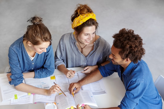 Top view of three students sitting at table surrounded with books and copybooks, discussing something with great interest, having happy expression. Brainstroming, team work and education concept