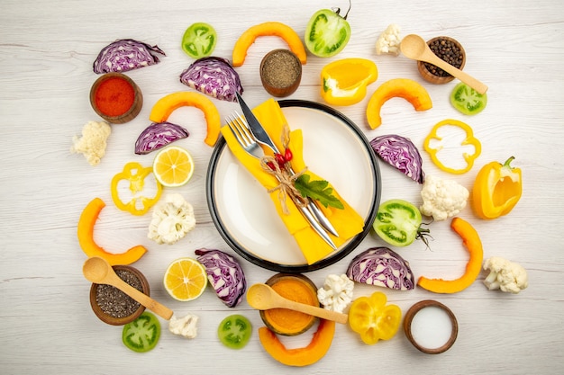 Top view tied knife and fork with yellow napkin on white platter cut vegetables red cabbage pumpkin cauliflower yellow bell pepper spices in small bowls on white table