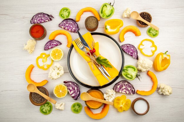 Top view tied knife and fork with yellow napkin on white platter cut vegetables red cabbage pumpkin cauliflower yellow bell pepper spices in small bowls on white table