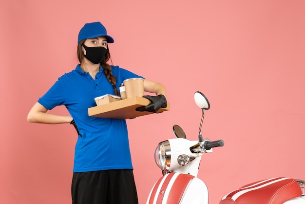 パステル ピーチ色のコーヒーの小さなケーキを保持しているオートバイの横に立っている医療マスク手袋を着て疲れた宅配便の女の子のトップ ビュー