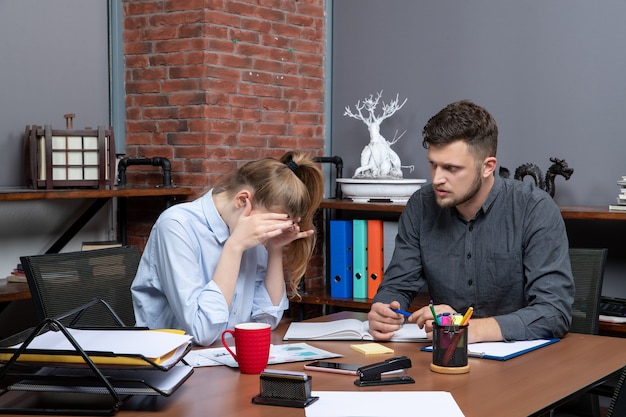 Top view of tired young female worker and her male co-worker sitting at the table in office enviroment