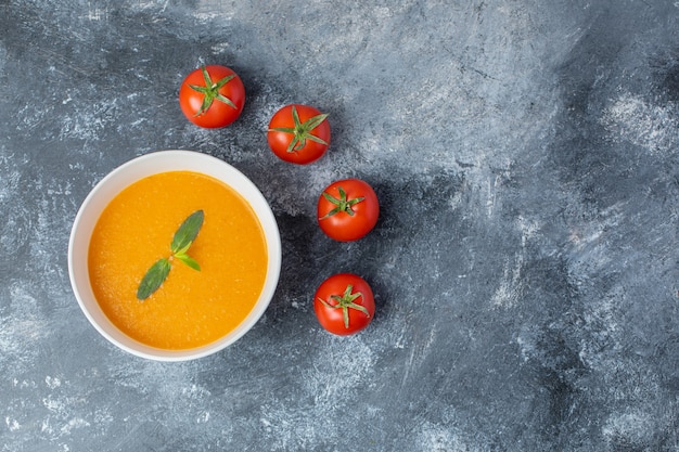 Top view of Tomato soup in white ceramic bowl with fresh tomatoes on grey table. 