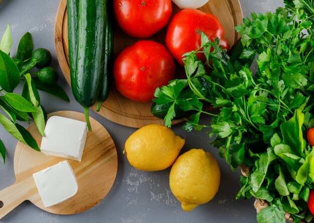 Top view tomatoes in cutting board with lemon, cucumber, cheese, greens on gray surface