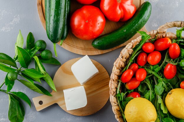 Top view tomatoes in cutting board with lemon, cucumber, cheese, greens on gray surface