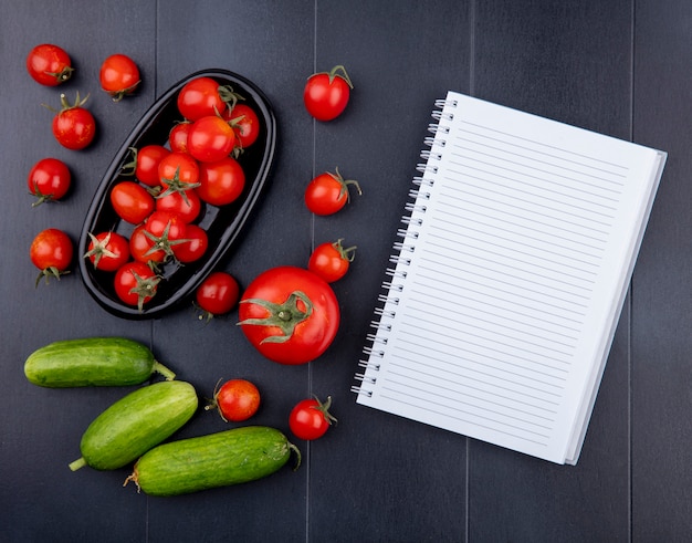 Top view of tomatoes in plate with cucumbers and note pad on black surface