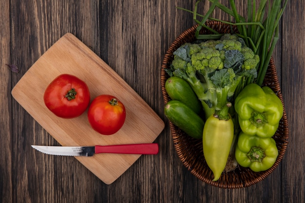 Top view tomatoes with a knife on a cutting board and cucumbers  green peppers  broccoli and green onions in a basket on a wooden background