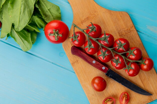 Top view of tomatoes with knife on cutting board and spinach on blue surface