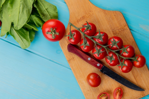 Вид сверху помидоры с ножом на разделочной доске и шпинатом на синей поверхности