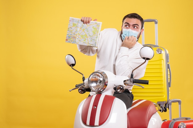 Вид сверху на концепцию путешествия с думающим парнем в медицинской маске, стоящим возле мотоцикла с желтым чемоданом на нем и держащим карту