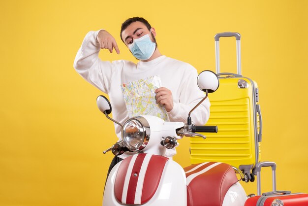 Вид сверху на концепцию поездки с уверенным в себе парнем в медицинской маске, стоящим возле мотоцикла с желтым чемоданом на нем и указывающим карту