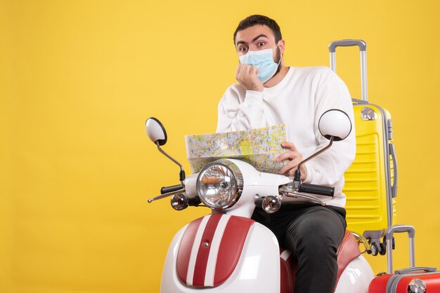 Vista dall'alto del concetto di viaggio con ragazzo confuso in maschera medica seduto su moto con valigia gialla su di esso e tenendo la mappa and