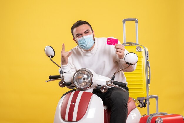 Top view of trip concept with emotional guy in medical mask sitting on motorcycle with yellow suitcase on it and holding bank card