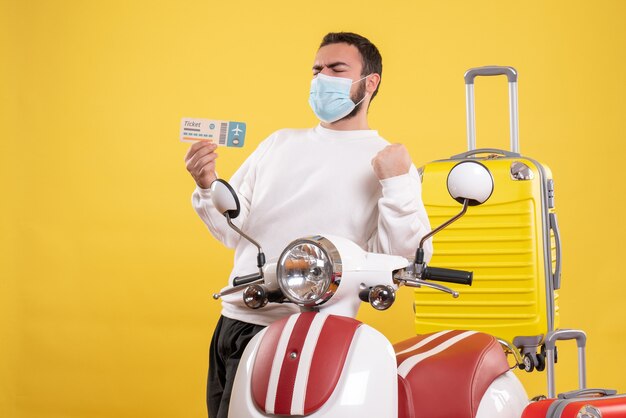 Top view of trip concept with proud guy in medical mask standing near motorcycle with yellow suitcase on it and holding ticket